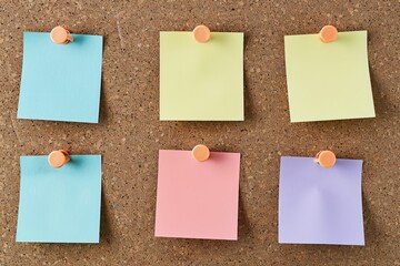 Six blank pastel-colored post-it notes are pinned neatly to a brown cork board
