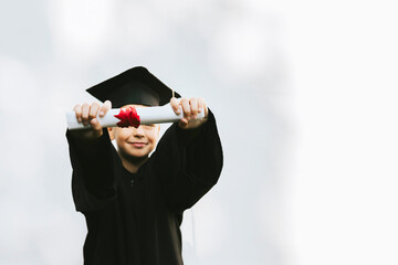 teenage boy in clothes of a graduate coat and cap celebrates high school or  junior year graduation on background of white wall with shadows and with diploma in hands, education and no school concept