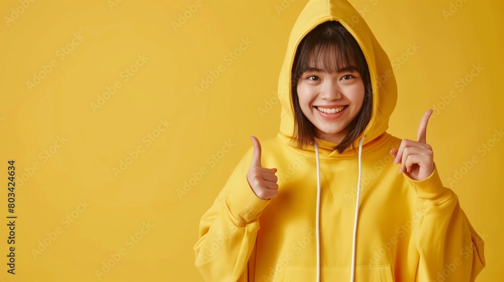 Poster Cheerful young Asian woman in a yellow hoodie giving thumbs up against a yellow background.