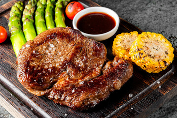 A beautifully grilled steak with asparagus, corn, tomatoes, and BBQ sauce, presented on a rustic wooden board