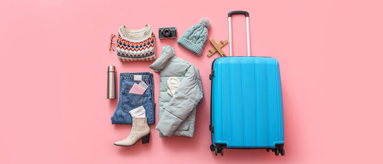 Suitcase with warm clothes, thermos and camera on pink background. Winter vacation concept