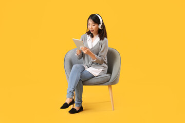 Beautiful Asian woman with headphones using tablet computer in grey armchair on yellow background
