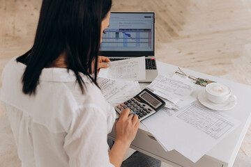 Back view of brunette woman calculates utility expenses on laptop and calculator, engaged in online...