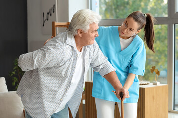 Young nurse helping senior man with stick to walk at home