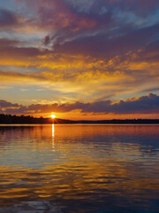 Illustrate the mesmerizing sight of a stunning sunset painting the horizon above a calm lake, its vibrant colors mirrored in the shimmering water below.