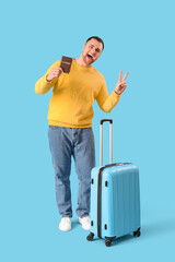 Male tourist with suitcase and passport showing victory gesture on blue background