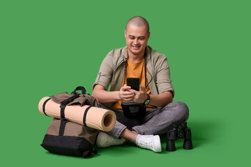 Young male tourist with backpack, binoculars and mobile phone sitting on green background