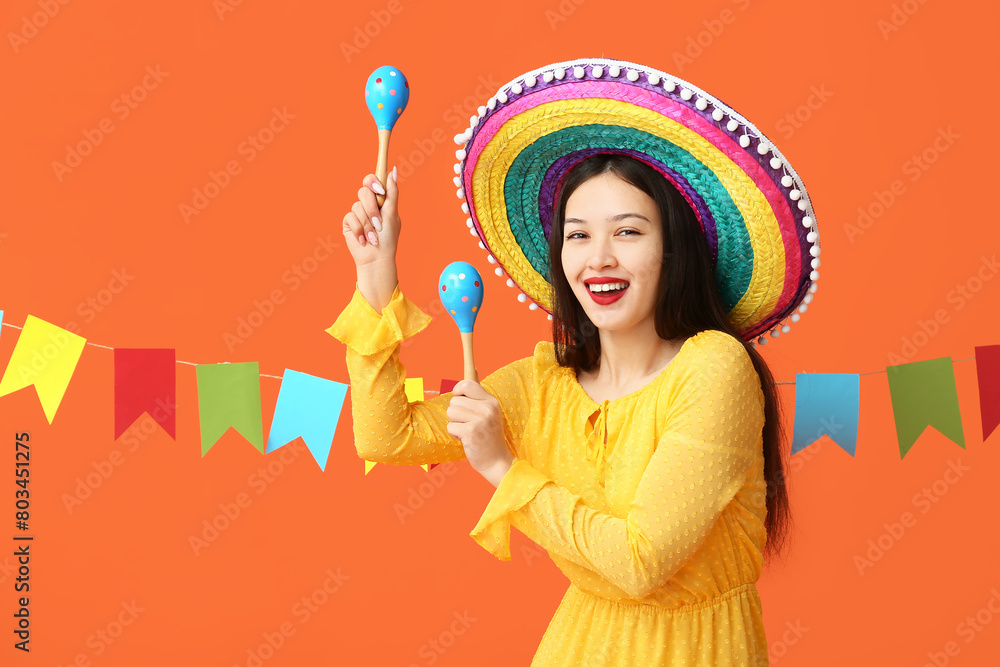 Wall mural Happy young woman in Mexican sombrero hat, with maracas and festive garland on orange background