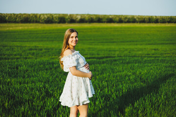 A smiling pregnant woman in a white light dress walks in a green spring garden. A woman is...