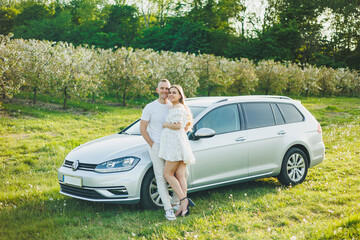 Happy married couple, pregnancy, man and woman in love walking in a blossoming apple orchard. A couple in love against the background of their car in nature.