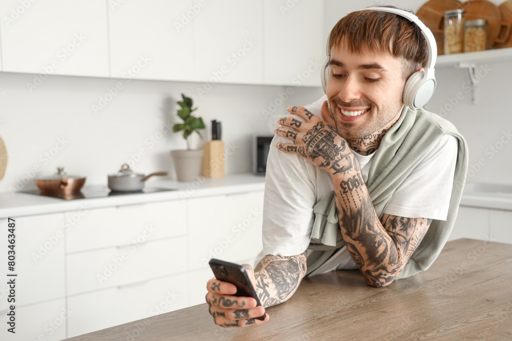 Poster young tattooed man with headphones and mobile phone listening to music at table in kitchen