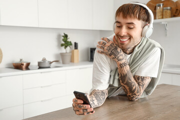 Young tattooed man with headphones and mobile phone listening to music at table in kitchen