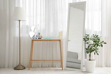 Floor lamp, mirror, houseplant and dressing table with set of decorative cosmetics near window