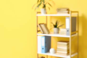 Bookcase with books and houseplants near orange wall in room, blurred view