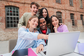 Group of multiracial teenage students smiling using a laptop to search information for the high school home works. Classmates studying for a exam watching the lesson on a notebook computer at campus