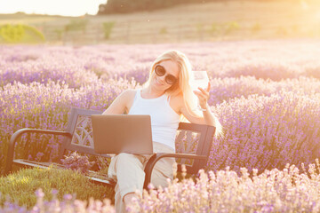 Lavender field. Woman sitting on bench among lavender flowers and working on laptop computer with...