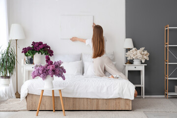 Young woman hanging picture on white wall near vases of lilacs in bedroom