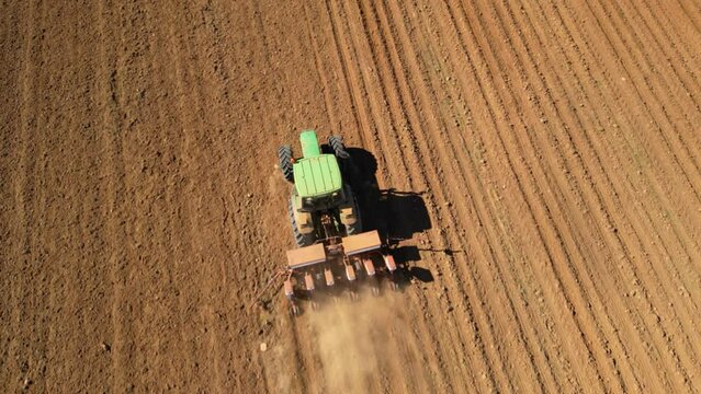 Aerial tracking high angle shot of tractor seeding a field with a seed drill in the mountains on a sunny spring day. Planter towed by a machine along a rural farmland. Ecologic farming concept.
