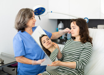 Female client looking at mirror while qualified beautician examining her face skin after procedure in medical cosmetology office.