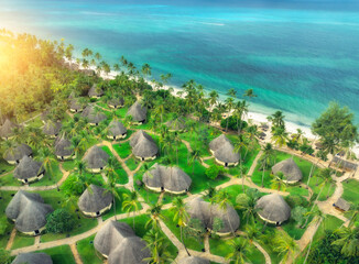 Aerial view of green palms, bungalows, grass, sandy beach, blue ocean at sunset in summer. Top view of wooden houses, sea with azure water, palms. Luxury resort in Zanzibar. Tropical background