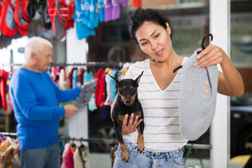 Asian woman choosing new clothing for her dog in pet shop. Old man making purchases in background.