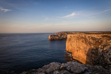 Landscape photo of dramatic, steep orange cliffs by the atlantic at sunset. Shot in Farol fo Cabo de Sao Vincente near Sagres, Portugal.