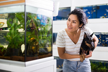 Positive asian woman with dog in her arms choosing aquarium fish at pet shop