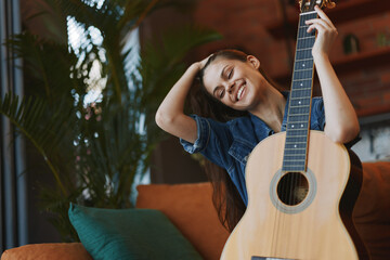 Young woman sitting on couch with acoustic guitar covering her face in cozy living room setting
