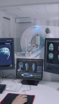 Vertical shot Female radiologist sits in control room and watches at monitors with displayed brain scans results. Professional doctor controls MRI or CT or PET scan with patient