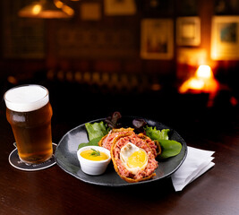 scotch egg with green salad and yellow mustard on plate with blurred background and half pint of craft beer