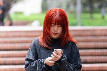 Young Korean woman with red hair deeply engrossed in using her smartphone outdoors