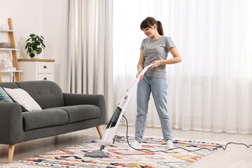 Happy young housewife vacuuming rug at home
