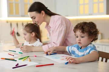 Mother and her little children drawing with colorful markers at table in kitchen