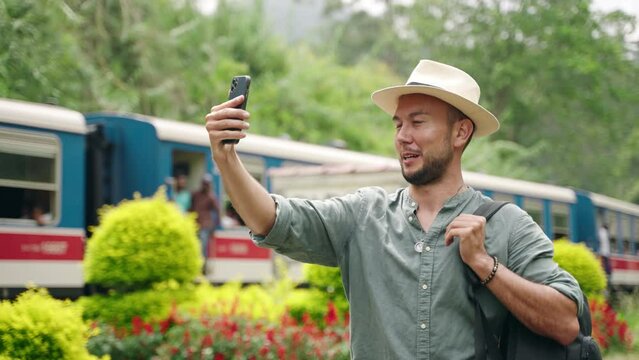 Asian male travel vlogger records selfie video at tropical train station. Content creator in hat captures journey, rich greenery, colorful train. Backpacking trip vlog, tourist explores. Slow motion.