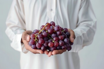 Saudi Gulf Arab man wearing a shemagh and white traditional dress is holding grape in hand on white...