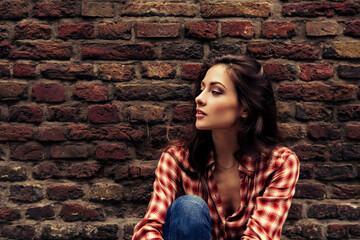 Beautiful brunette woman with long hair sitting near the old brick wall building background in...