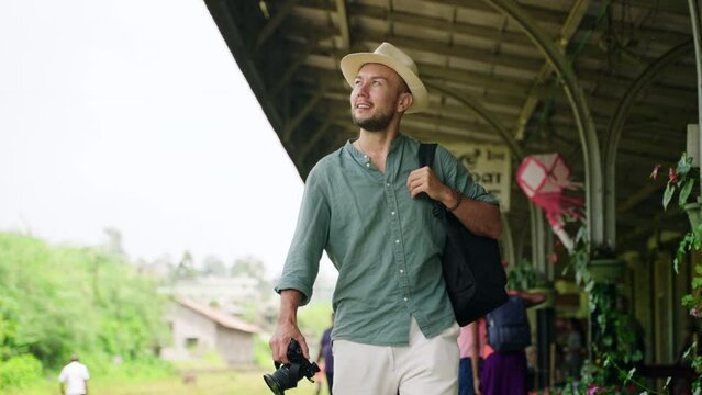 Asian male tourist explores Haputale railway station with camera and backpack. Solo traveler in hat enjoys scenic train depot in Sri Lanka. Adventure, culture, wanderlust blend as man journeys alone.