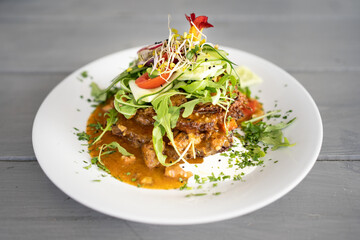 An elegantly plated dish featuring crispy cutlets topped with a vibrant salad of fresh greens, sprouts, and flowers, served on a circular white plate with a sauce and herb garnish.