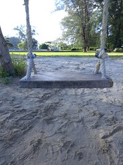 A Rustic Wooden Swing Suspended by Ropes on a Sandy Beach Playground, Beckoning for Play Under a Bright Daytime Sky with Distant Trees
