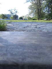 a Rustic Wooden Plank Across a Sandy Expanse Towards a Green Park Landscape with Distant Trees Under a Bright Daytime Sky