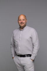 A studio portrait of a bald man in a light grey shirt, looking directly at the camera with a slight...