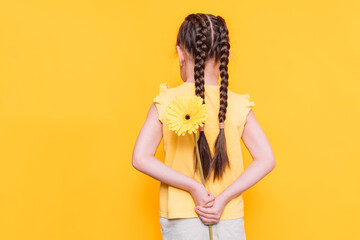 Little girl holding a beautiful flower behind her back against a yellow background.
