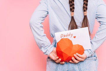 Little girl holding a drawn card and a heart behind her back for Father's Day holiday in front of a pink background.