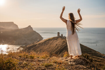 A woman in a white dress stands on a rocky hill overlooking the ocean. She is smiling and she is...