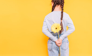 Child holding a yellow flower behind her back against a yellow background.