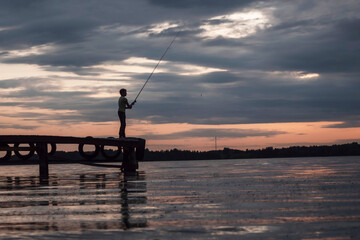 A man with a fishing rod on a wooden bridge against the background of sunset.