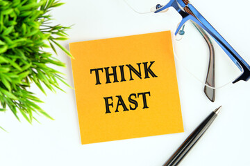 THINK FAST words on a yellow sticker on a light background next to glasses, a pen and a green plant