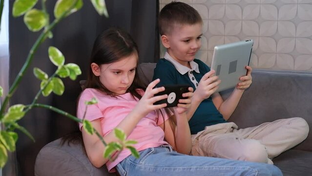 Two children boy and girl, play games or watch informational video text on Internet on mobile phones and tablet computers at home on sofa. Leisure, children, digital technology and people concept.