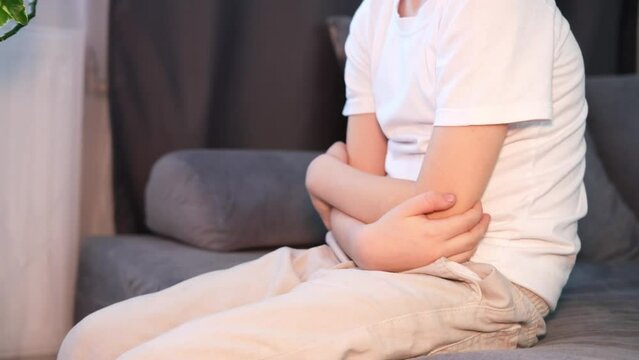 Little boy suffers from nausea in the living room. Child suffers from abdominal pain while sitting on sofa at home. Child holds his hands on his stomach. Diarrhea or gastroenteritis. Health problems.	