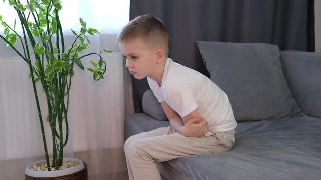 Little boy suffers from nausea in the living room. Child suffers from abdominal pain while sitting on sofa at home. Child holds his hands on his stomach. Diarrhea or gastroenteritis. Health problems.	